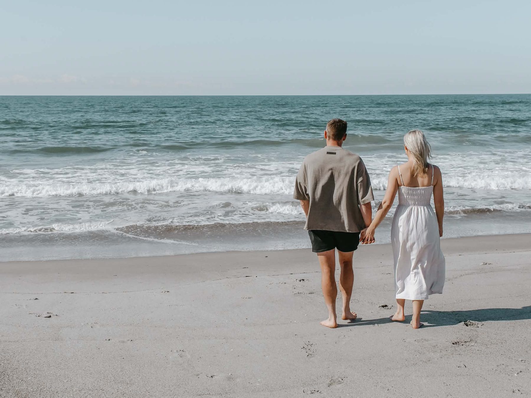 Lacy and Josh walking hand in hand towards the ocean on a wide sandy beach, seen from behind