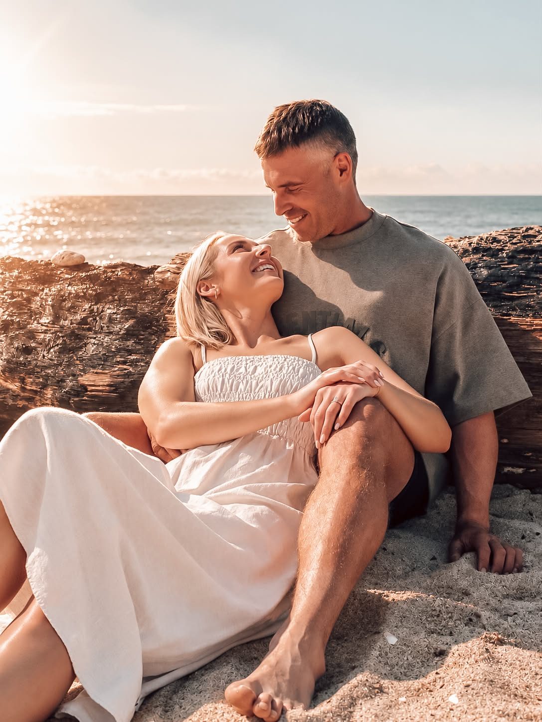 Lacy and Josh sitting together on the sand at golden hour, smiling at each other